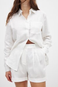 Hvid linned skjorte med knaplukning og lomme, parret med matchende plisserede shorts. Begge dele har et let, tekstureret udseende.
