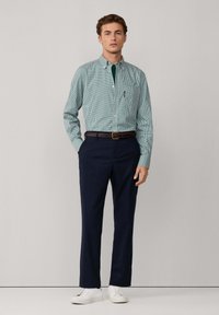 Joven de pie, vistiendo una camisa de cuadros verde con botones, pantalones de rayas azul marino, cinturón marrón y zapatillas blancas contra un fondo liso.