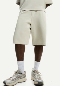 Beige shorts with a relaxed fit, side pockets, and a smooth texture. Paired with light socks and multi-colored athletic shoes.