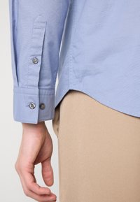 Camisa de manga longa azul claro com punho abotoado e textura subtil; usada sobre um tecido bege, mostrando parte da mão e a bainha curva da camisa.