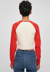 Person mit kurzen lockigen Haaren, der ein weißes und rotes Langarmshirt und eine blaue Jeans trägt und mit dem Rücken zu einem schlichten Hintergrund steht.
