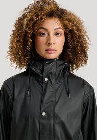 Mustavedenkestävä takki korkealla kauluksella, etupainikkeilla, säädettävällä nyöriyllä ja sileällä pinnalla. Mallilla on kiharat hiukset ja neutraali tausta.