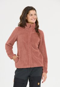 Ruskon värinen fleece-takki, jossa on teksturoitu sahalaitakuviotus, korkea kaulus ja etuvetoketju. Ominaisuuksiin kuuluvat kaksi sivutaskua ja pitkät hihat.