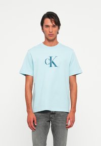 Muškarac odjeven u svijetloplavu majicu s logom Calvin Klein i izblijedjele sive traperice, stoji naslonjen na jednostavnu bijelu pozadinu, suočen prema naprijed s neutralnim izrazom lica.