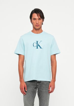Hombre vestido con una camiseta de Calvin Klein de color azul claro y jeans gris desgastados, de pie contra un fondo blanco liso, mirando de frente con expresión neutral.