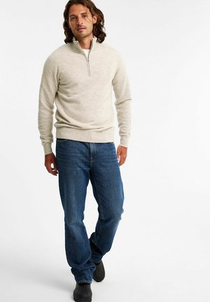 Lys beige zip-neck trøje med ribbede manchetter og kant, parret med mørkeblå afslappede jeans og sorte sko, der står foran en neutral baggrund.