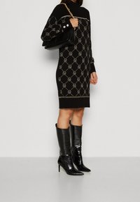 Kvinna iklädd en svart stickad klänning med ett geometriskt mönster, svarta knähöga klack stövlar med spetsig tå, och bär en svart handväska med en guldkedjestropp.