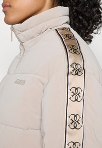 Nærbilde av en person som har på seg en beige puffjakke med høy krage og et merke tape med et gjentakende svart logo på skulderen.
