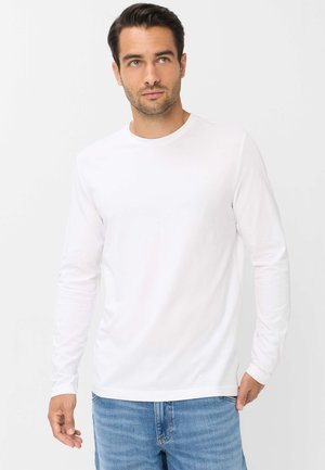 Ung mand med kort mørkt hår iført en ensfarvet hvid langærmet skjorte og blå jeans, stående mod en hvid baggrund.