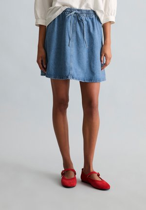 Personne portant une jupe en denim bleu à taille coulissante, un chemisier blanc et des ballerines rouges style Mary Jane, debout sur un sol gris clair.