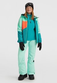 Skikleding met een teal kleurige jas met oranje en aqua accenten, gecombineerd met mintgroene broek en zwarte handschoenen, geschikt voor wintersport.