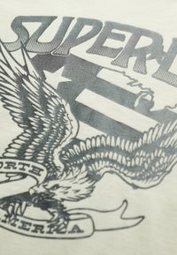 Beżowa koszulka z czarnym wzorem graficznym przedstawiającym orła oraz banerowym tekstem „NORTH AMERIKA”, nałożonym na wzór w paski flagi.