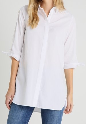 Woman wearing a long white button-up shirt with 3/4 sleeves tied at the elbows and blue jeans, standing against a plain background.