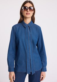 Kobieta nosząca oversize'owe okulary w tortoise shell, niebieską dżinsową koszulę z długim rękawem i detalem w postaci wiązania, a także pasujące dżinsowe spodnie.