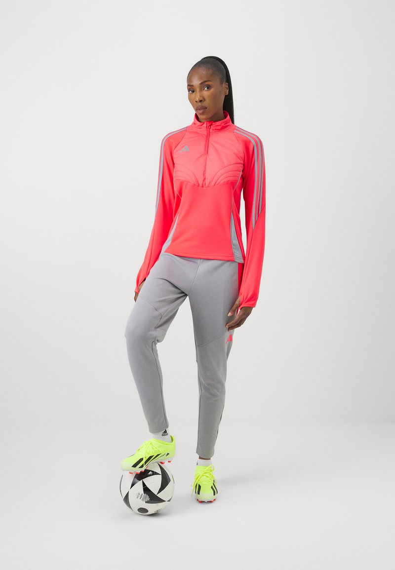Rosa hochgeschlossener Sportjacke mit grauen Akzenten, kombiniert mit hellgrauen Hosen und leuchtend gelben Fußballschuhen, stehend auf einem Fußball.