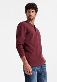 Man med lockigt hår som bär en vinröd långärmad tröja och blå jeans, tittar åt vänster mot en vit bakgrund.