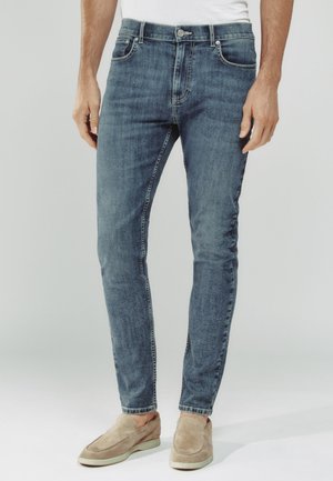 Mann trägt slim-fit ausgewaschene blaue Jeans, weißes T-Shirt und beige Wildleder-Slipper vor einfachem hellem Hintergrund.