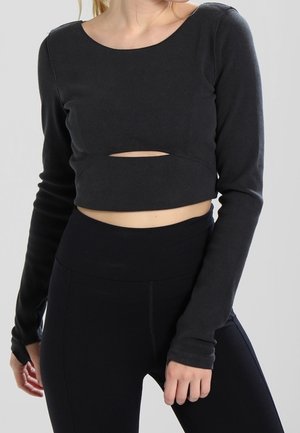 Vrouw draagt een zwarte cropped top met lange mouwen en een buikuitsnede en zwarte leggings met hoge taille, staand tegen een effen achtergrond.
