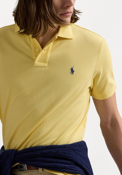 Tricou polo galben cu un finisaj texturat, având un logo brodat albastru. Un pulover bleumarin este legat la talie.