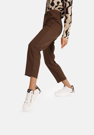 Pantaloni marroni sartoriali con dettagli sulle cuciture laterali abbinati a sneaker bianche con accenti dorati e finitura testurizzata.