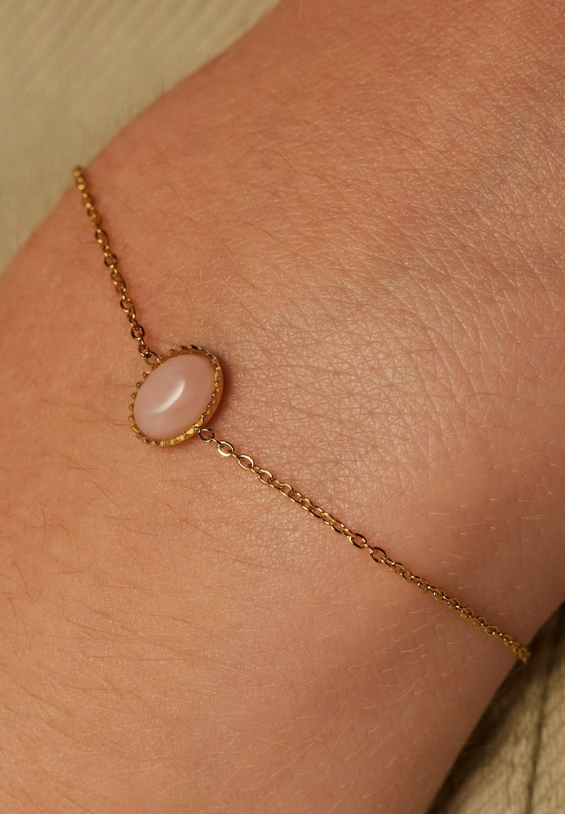 Gold chain bracelet featuring an oval, pink stone centerpiece with a scalloped bezel, resting on a light skin tone.