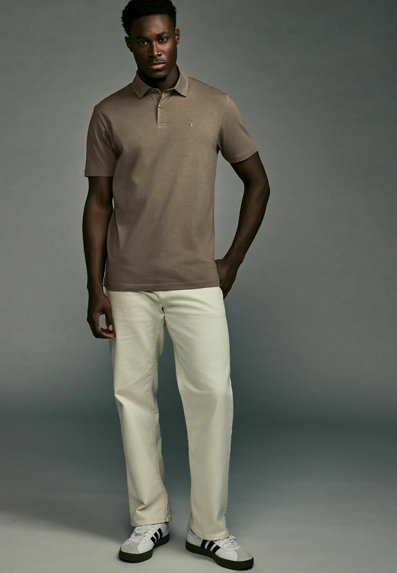 Polo-Shirt aus leichtem braunen Baumwollstoff, mit kurzen Ärmeln, Kragen und Logo; kombiniert mit cremefarbener Hose und schwarz-weißen Sneakern.