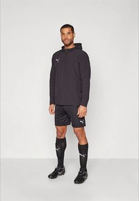 Vīrietis, kas ģērbies melnā Puma sporta jakā, šortos, zeķēs un futbolu apavos, stāvot pret vienkāršu gaismas fonu.