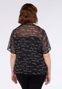 Femeie cu părul scurt de culoare brună, îmbrăcată cu o bluză subțire neagră cu text alb și bej, și pantaloni negri, stând cu spatele întors spre cameră.