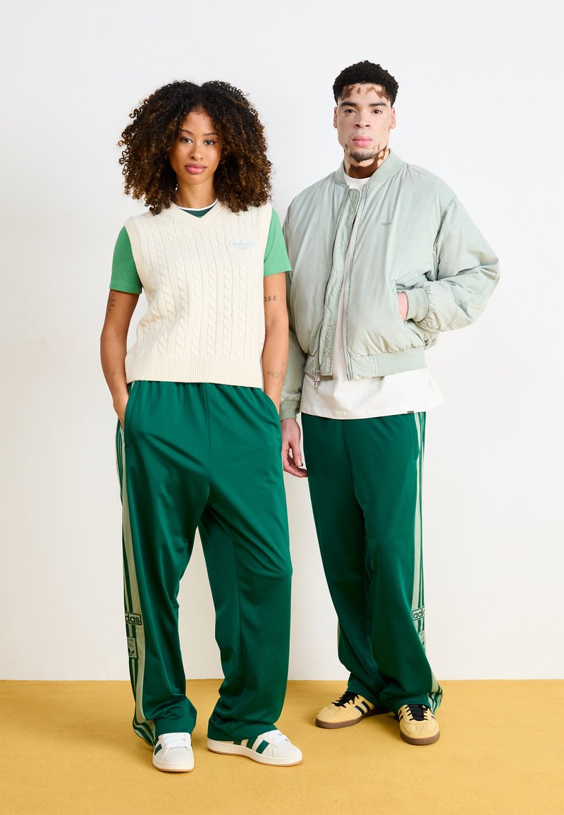 Dos personas están de pie una al lado de la otra. Una lleva un chaleco de punto en color crema sobre una camisa verde y pantalones de chándal verdes. La otra lleva una chaqueta bomber verde claro y pantalones de chándal verdes a juego.