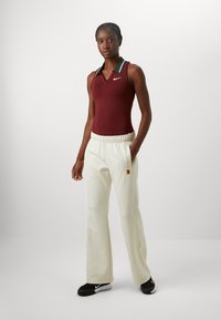 Top sin mangas color burdeos con cuello negro y aqua, combinado con pantalones de pierna ancha color crema, que cuentan con un bolsillo y un detalle de logo. Calzado negro.