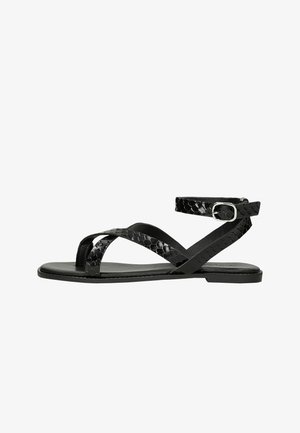Schwarze flache Sandale mit dünnen kreuzweise verlaufenden Riemen und einem Knöchelriemen mit silberner Schnalle, strukturiert mit einem dezenten Schlangenmuster.