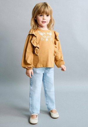 Mlada djevojka sa plavom kosom do ramena, odjevena u žutu cvjetnu bluzu s naborima, svijetloplave traperice i bijele baletanke, stoji na sivoj pozadini.