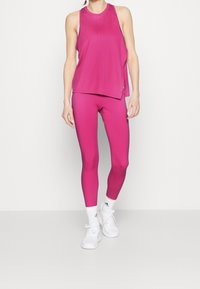 Fuksjowy bezrękawnik sportowy w połączeniu z dopasowanymi, obcisłymi legginsami, wykonanymi z gładkiego materiału, uzupełniony białymi butami sportowymi.