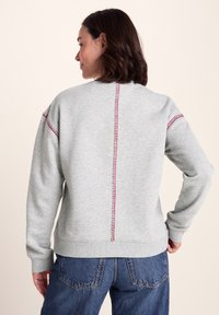 Gray sweatshirt with a red accent stitch along the back seam. Features a relaxed fit and ribbed cuffs. Smooth cotton fabric.