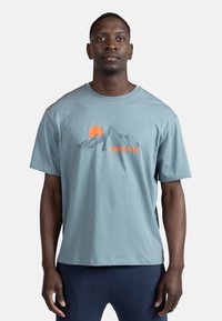 Lysblå bomulds t-shirt med korte ærmer, der har et bjergmotiv og en orange solillustration over teksten "ROSSIGNOL" i orange.