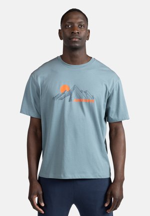 Camiseta de algodón azul claro con mangas cortas, que presenta un gráfico de montaña y una ilustración de sol naranja sobre el texto "ROSSIGNOL" en naranja.