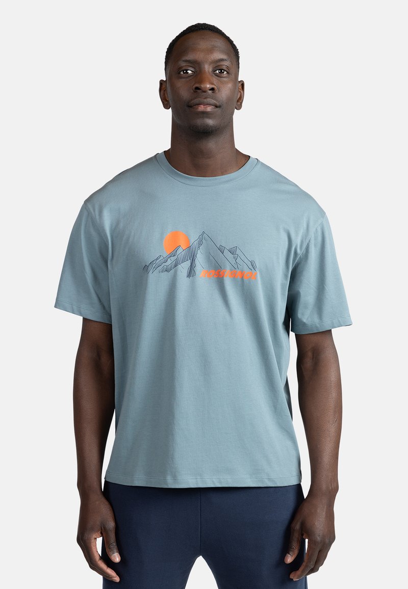 Lysblå bomulds t-shirt med korte ærmer, der har et bjergmotiv og en orange solillustration over teksten "ROSSIGNOL" i orange.