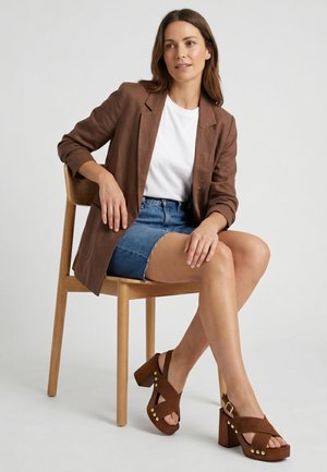 Frau sitzt auf einem Holzstuhl, trägt braunen Blazer, weißes Hemd, Jeansrock und braune Plateausandalen mit goldenen Nieten.