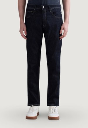 Person iført mørkeblå, lige jeans og hvide sneakers, stående mod en ensartet, lys baggrund.