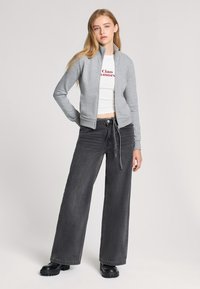 Felpa grigia con zip e colletto alto, indossata sopra una maglietta cropped bianca con testo rosso, abbinata a pantaloni neri a gamba larga e stivali neri chunky.