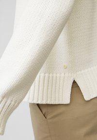 Primo piano di una manica e orlo di un maglione lavorato a maglia color crema, con una piccola apertura laterale, indossato sopra pantaloni color beige.