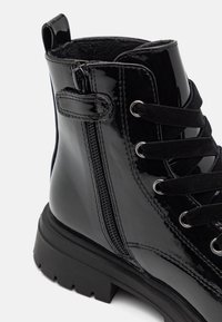 Botín de charol negro con cremallera lateral, cordones negros pasados por ojales de metal y suela de goma gruesa.