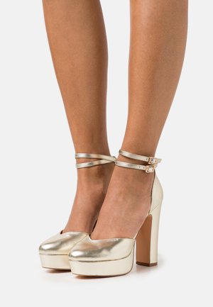 Chaussures à plateforme dorées à talons haut avec des brides de cheville. Présentent un bout arrondi, une texture lisse et un talon épais en forme carrée.