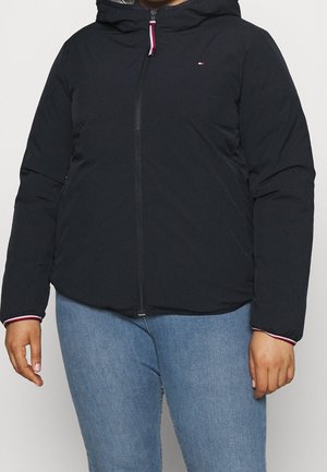 Person trägt eine schwarze Kapuzenjacke mit Reißverschluss und kleinem Logo auf der Brust sowie blaue Jeans, steht vor einem einfarbigen Hintergrund.