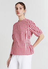 Giovane donna che indossa una blusa a maniche corte rosso e bianco a quadretti e pantaloni bianchi, in piedi con una mano sui fianchi, sfondo neutro.