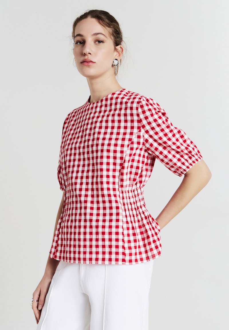 Giovane donna che indossa una blusa a maniche corte rosso e bianco a quadretti e pantaloni bianchi, in piedi con una mano sui fianchi, sfondo neutro.