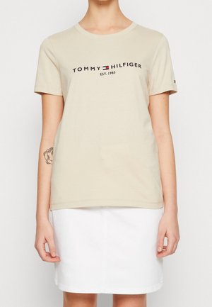 Personne portant un t-shirt beige Tommy Hilfiger avec logo et texte « est. 1985 », associé à une jupe blanche et un tatouage sur l'avant-bras gauche.