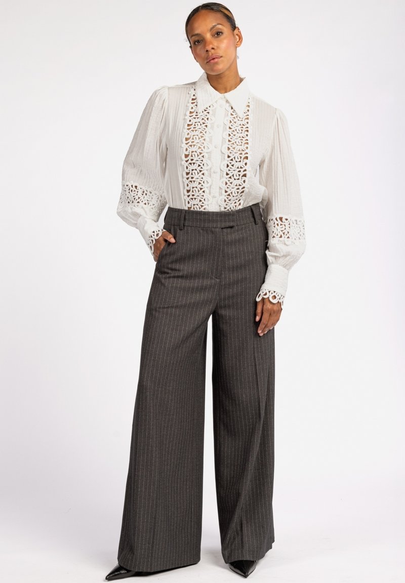 Witte blouse met kantdetails en knoopfront. Grijze gestreepte broek met wijde pijpen. Gladde stof, gestructureerde pasvorm en puntige schoenen.