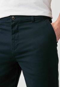 Tamsiai mėlynos chino kelnės su sklandžiu paviršiumi, turinčios mygtuko užsegimą, šonines kišenes ir pritaikytą siluetą. Minimalistinis dizainas be raštų.