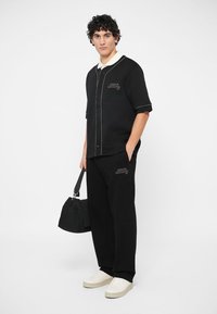 Camisa preta de mangas curtas com botões e detalhes brancos, combinada com calças pretas. Um pequeno logótipo é visível na camisa e nas calças. Ténis de cor clara.